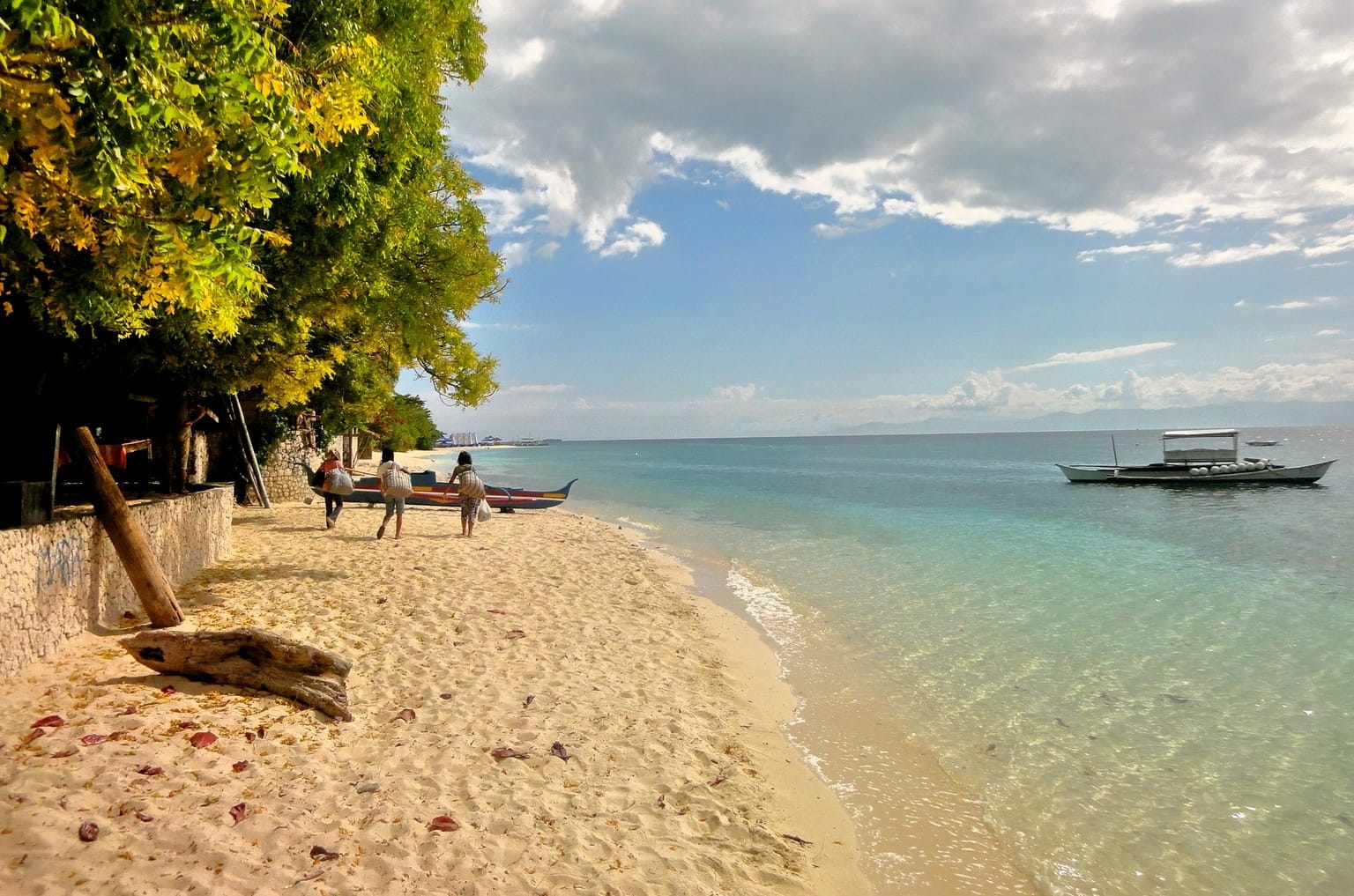 White sand and clear turquoise water at Basdaku White Beach in Moalboal, with trees along the shore and an outrigger boat moored offshore