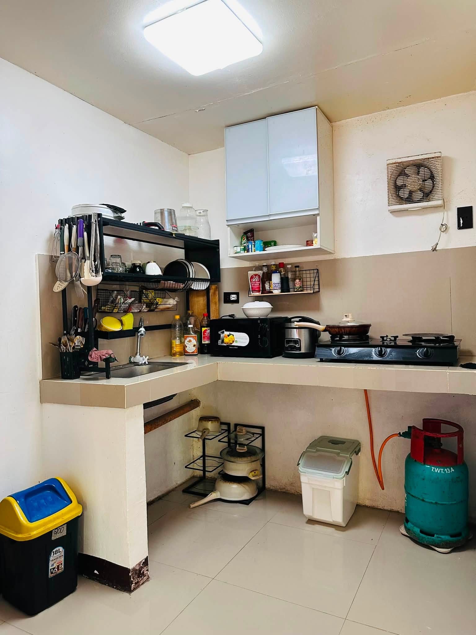 Kitchen with an L-shaped tiled counter, sink, utensil rack, microwave, rice cooker and a two-burner gas stove