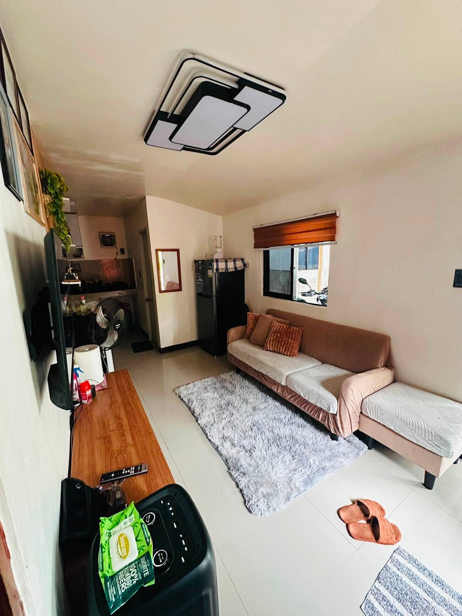 Living room seen from the TV wall, with the sofa, gray rug, refrigerator, a window with wooden blinds and the kitchen at the far end