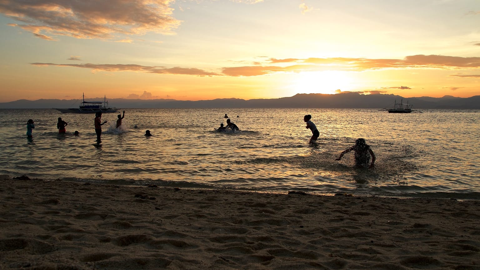 People swimming at Panagsama Beach at sunset, with outrigger boats on the water and the Negros coast in the distance
